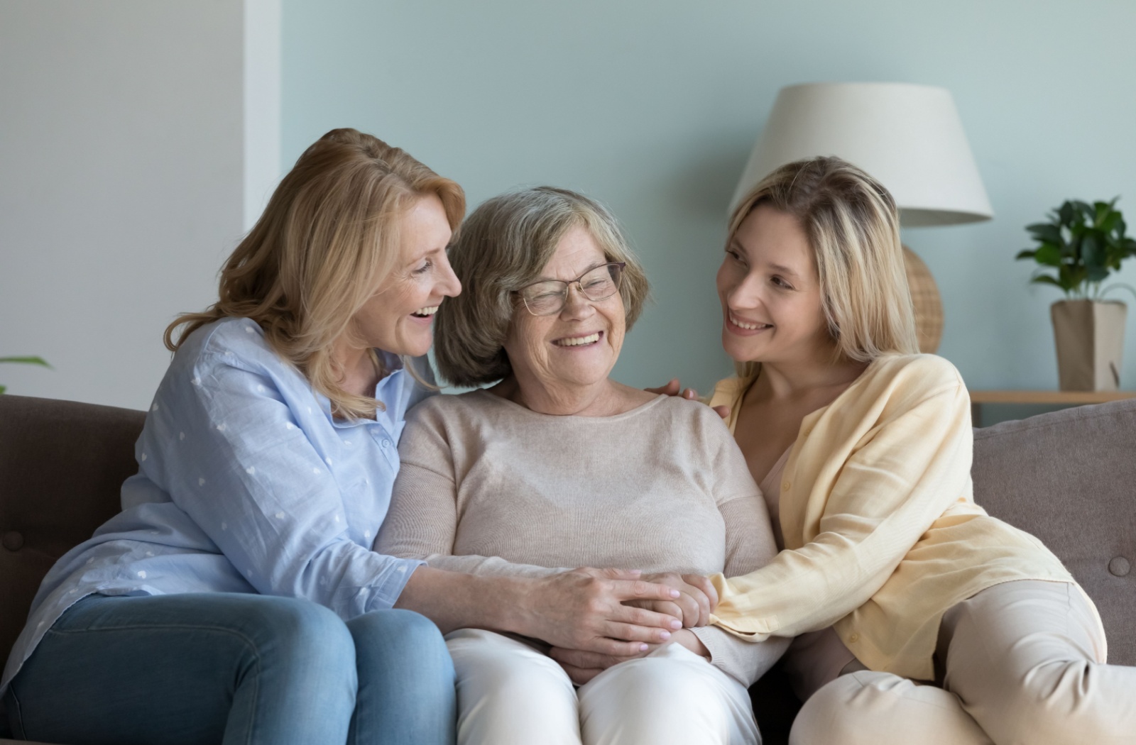 A grandmother in memory care being visited by two of her family members, holding hands with her daughter and granddaughter on the couch and smiling.
