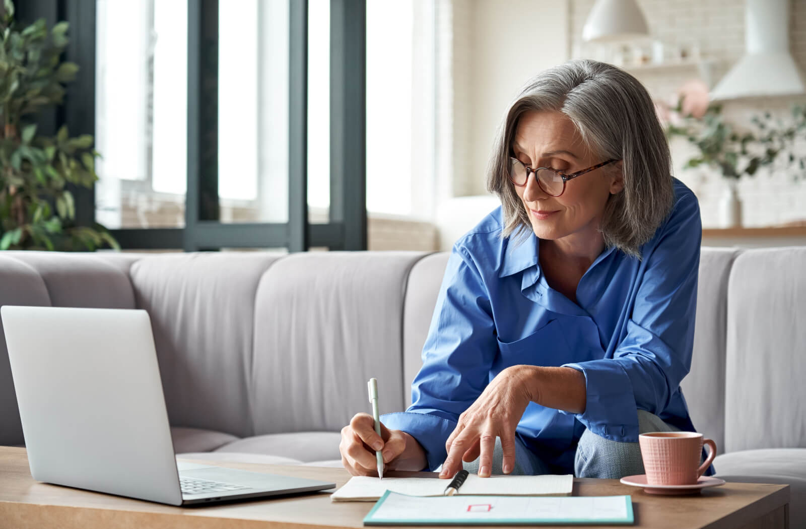 An older adult sitting on their couch in front of their laptop, writing notes in a notepad to test their memory.
