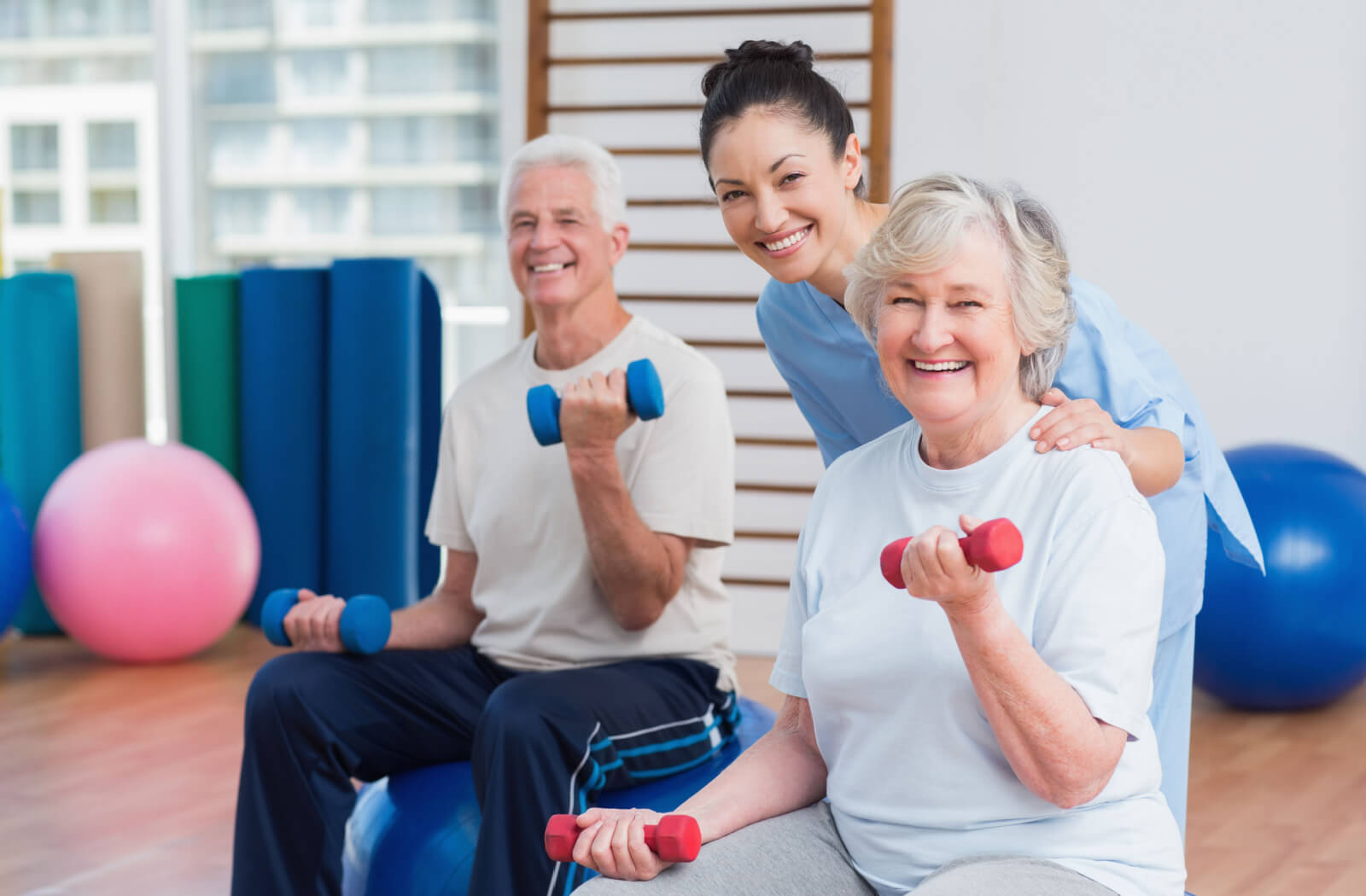 A caregiver supporting 2 seated older adults while they use small handheld weights to exercise.
