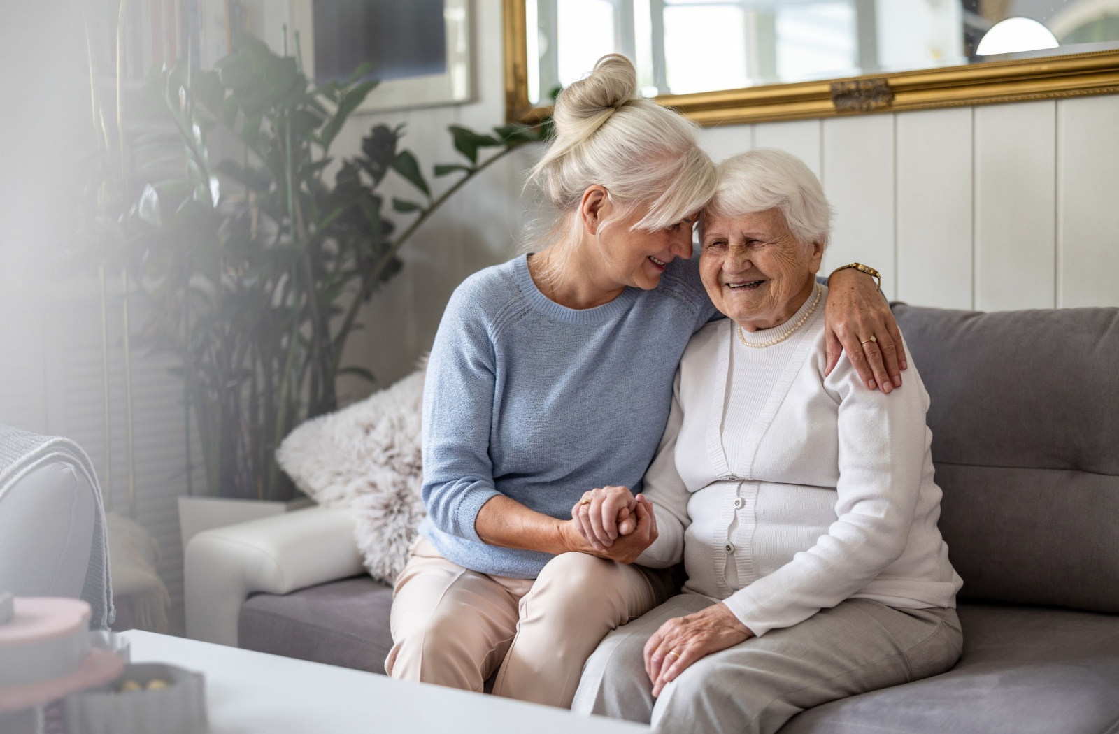 An adult and their parent smile together on the couch of a memory care community.