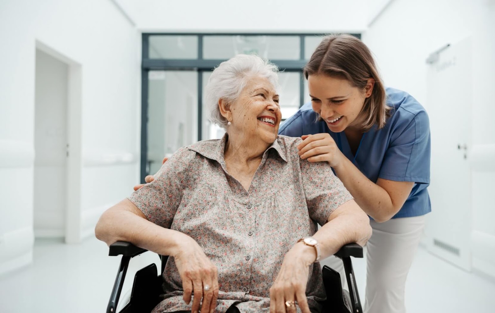 A nurse laughs along with a memory care resident while pushing their wheelchair down a hallway.