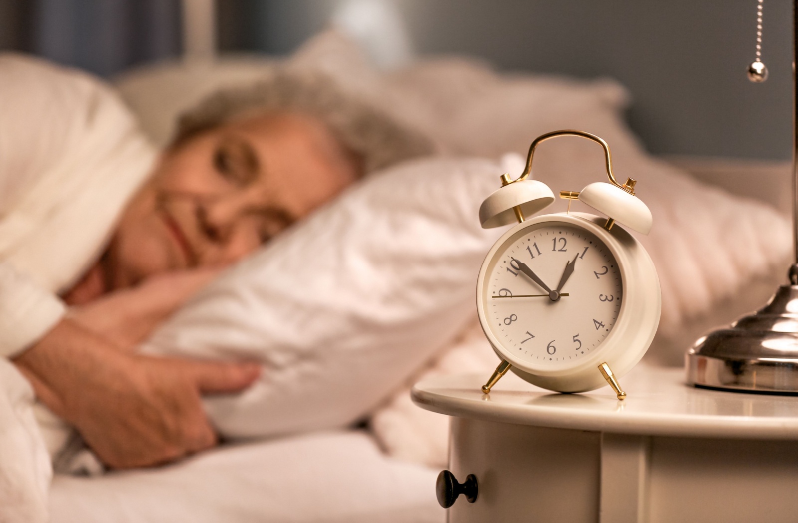 A close-up image of an alarm clock late at night while an older adult sleeps in the background.