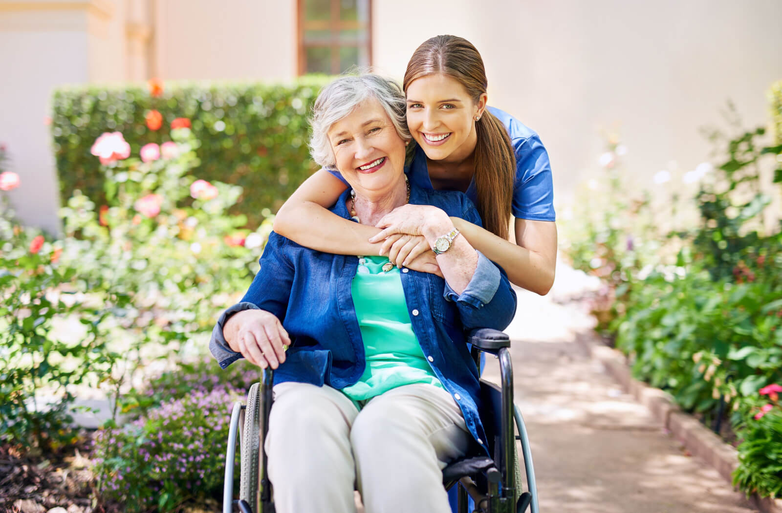 A caregiver hugs an older adult from behind while on a walk together in a garden.