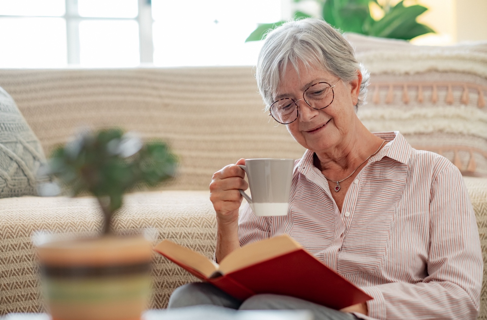 An older adult sips tea while reading a hardcover book and smiling in memory care