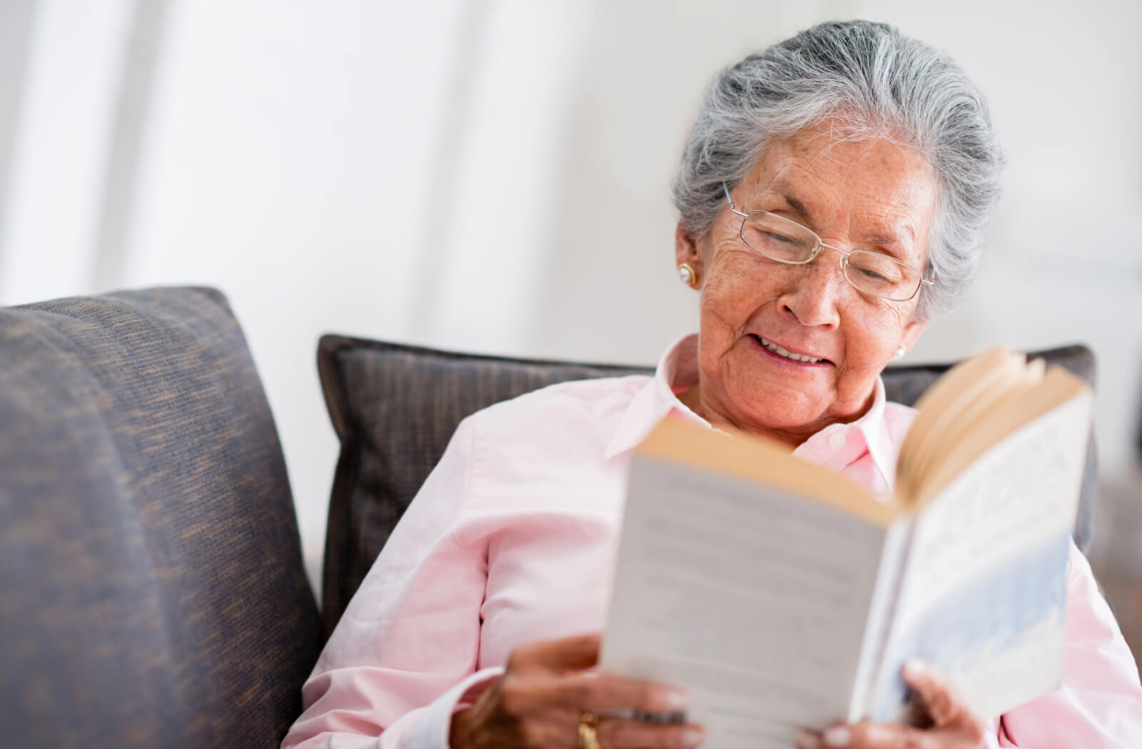 An older adult laughs while reading a paperback novel on their couch in memory care