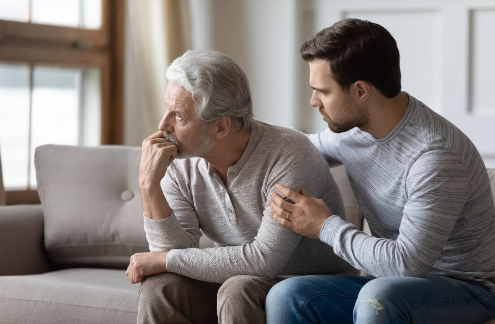 An adult child sits beside their older parent on the couch, rubbing their back and hugging them to reassure them about their recent memory loss
