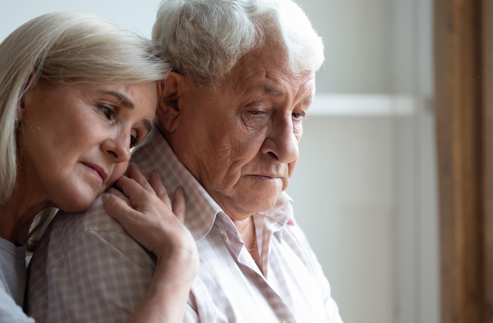 An older adult hugs their spouse from behind to support them, with both of their faces showing stress due to dementia

