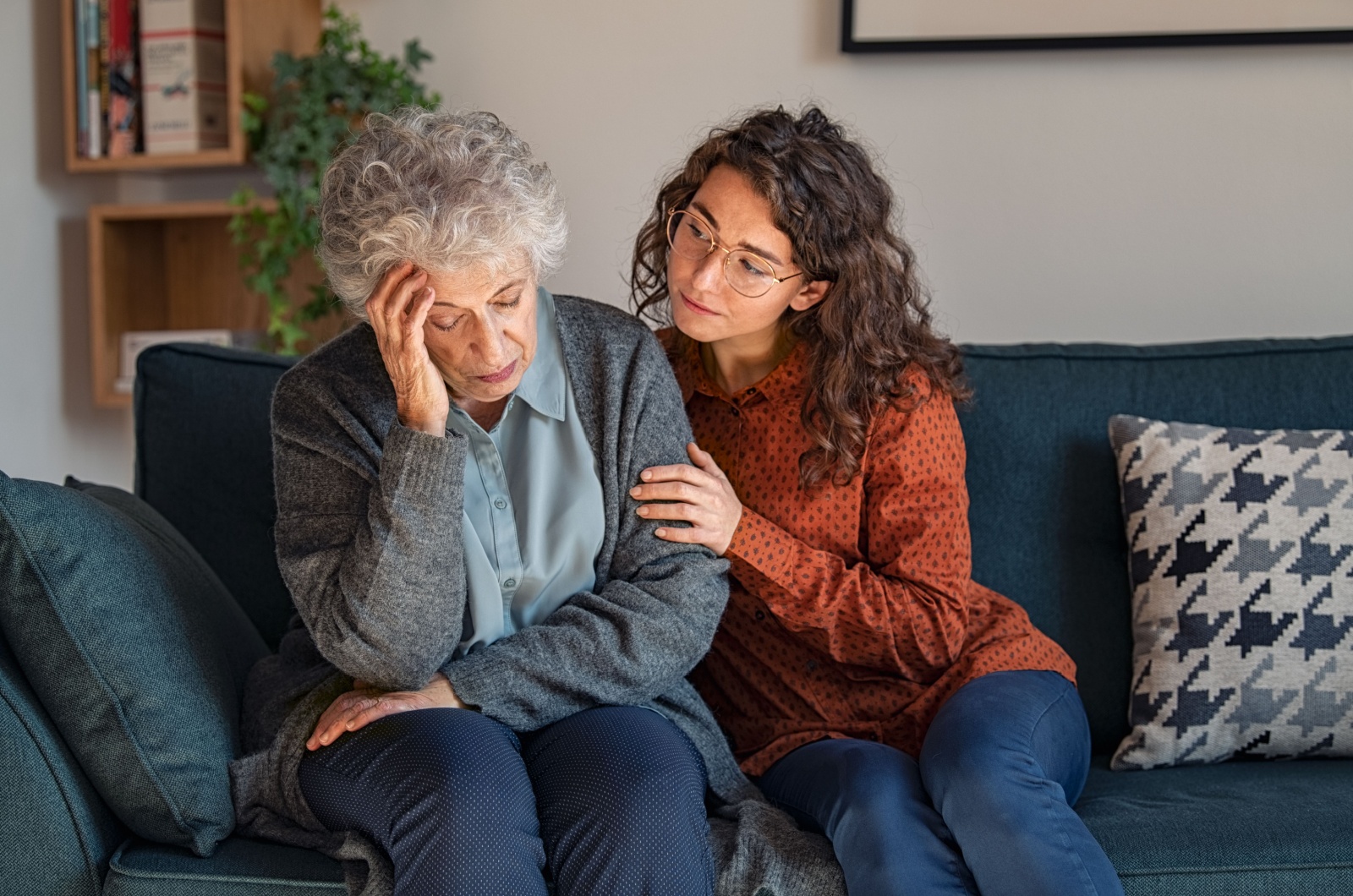 An adult child comforts their parent with dementia as they both sit on a blue couch.