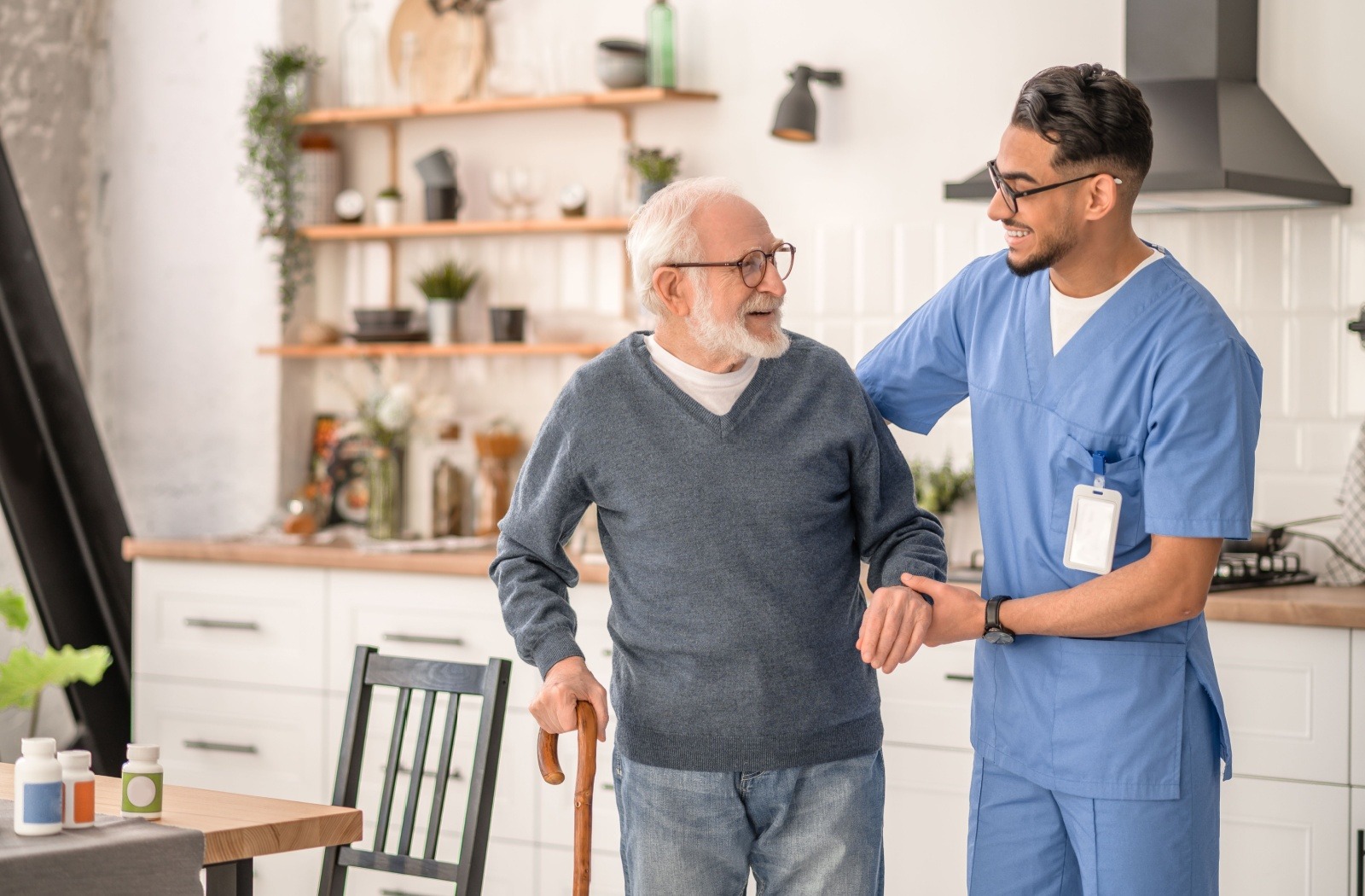 A caregiver in memory care assisted a smiling older adult as they walk through the kitchen together